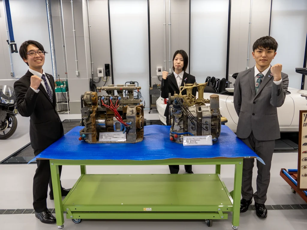 マツダ自動車整備専門学校（神戸）の実習場で、マツダの魂であるロータリーエンジンの横で力強いガッツポーズを決める、真新しいスーツ姿の第一期生（開拓者）たち